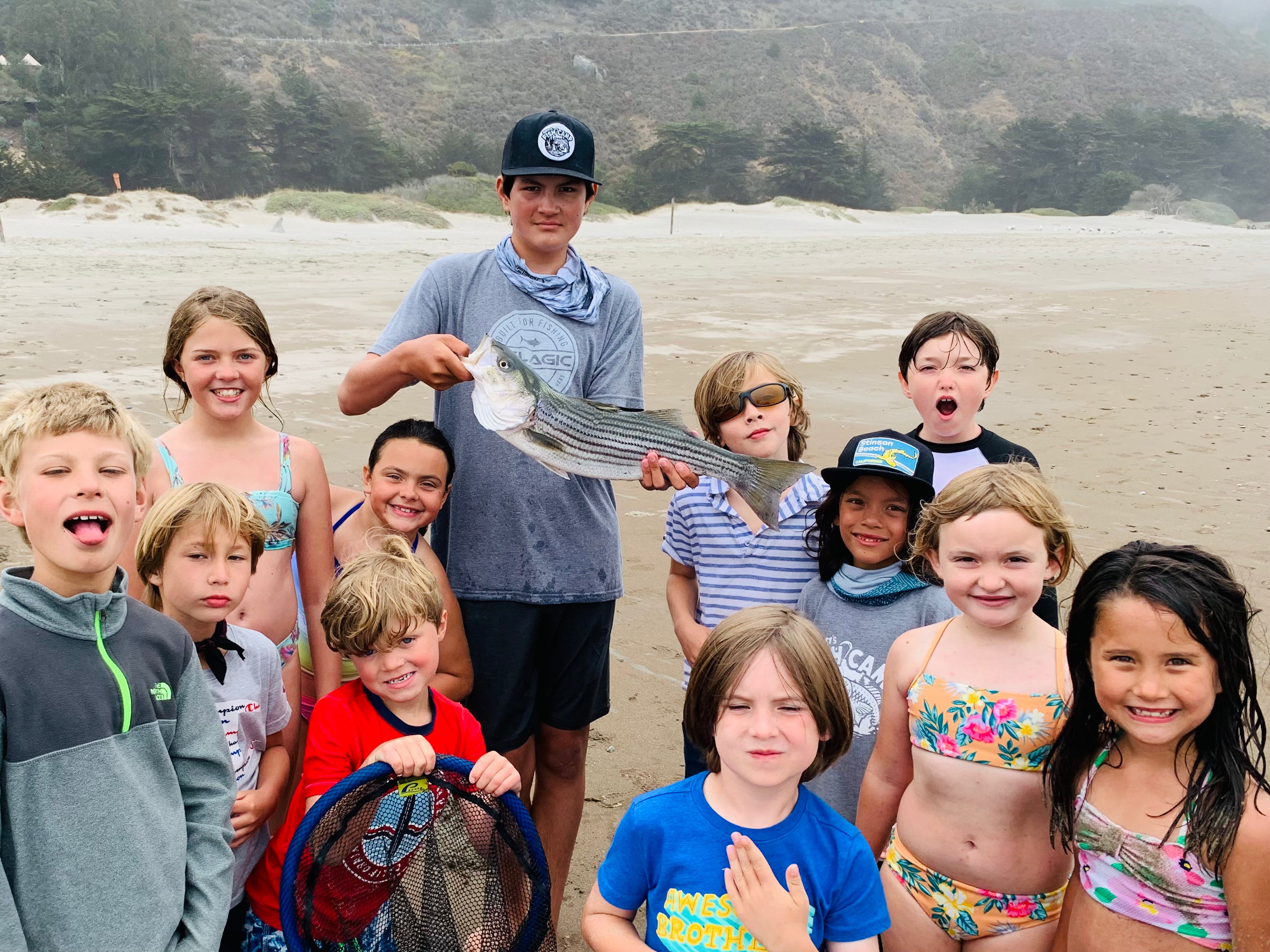 Children on a beach holding a fish, with a scenic background of trees and sand.