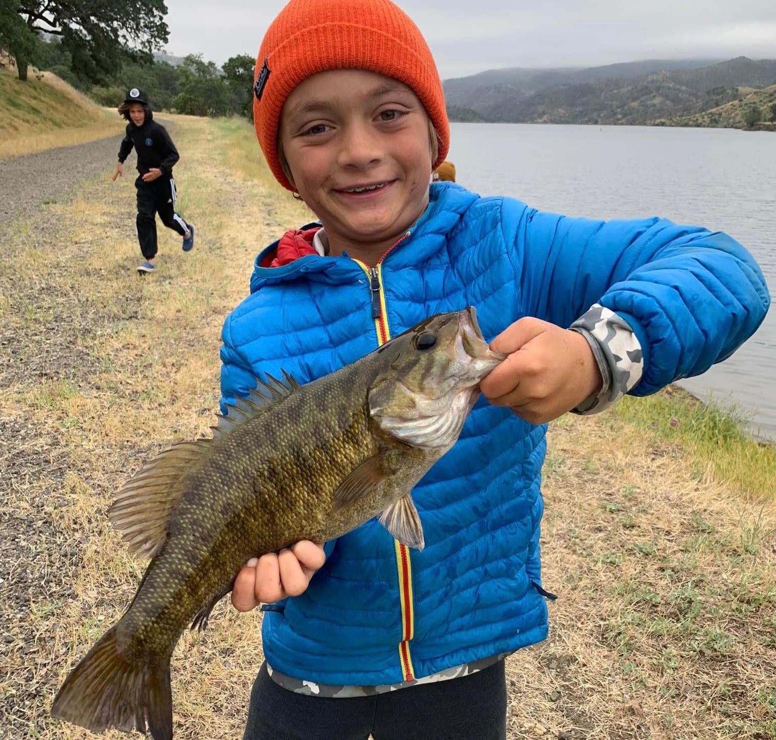 Child holding a fish by a lake with another person in the background